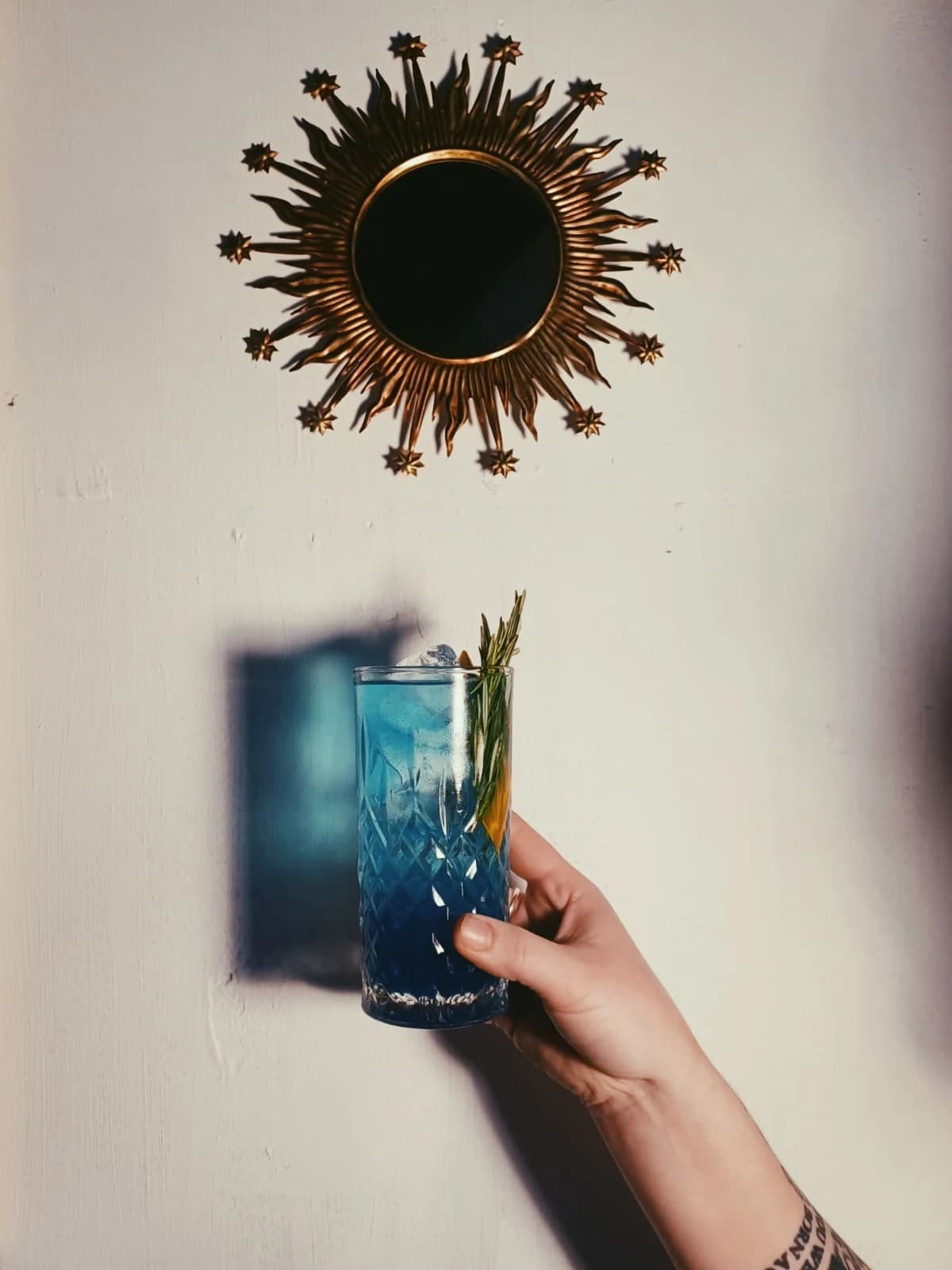 A blue cocktail held up beside a sunburst mirror